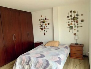 A bedroom with a large wooden wardrobe, a bed with a patterned bedspread, a nightstand with a lamp, and two wall art pieces. - Beautiful Suite in Cuenca (Cuenca)