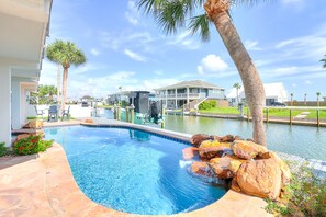 A swimming pool with a curved edge, surrounded by rocks and palm trees, with houses in the background. - Canal-front home with saltwater pool, pool spa, kayaks, dock, & casita (Rockport)