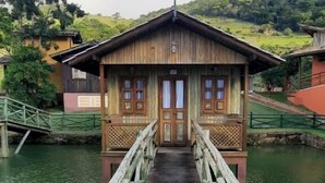 A wooden house with a wooden bridge leading to it, surrounded by water and greenery.