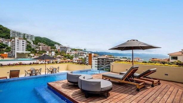 Un espace piscine aménagé sur le toit, avec des chaises longues et une table, offrant une vue sur le paysage urbain et les montagnes.