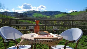 Une table recouverte d’une nappe tissée, deux chaises, un pain, une bouteille et deux verres de vin rouge, le tout disposé devant un paysage pittoresque de collines.
