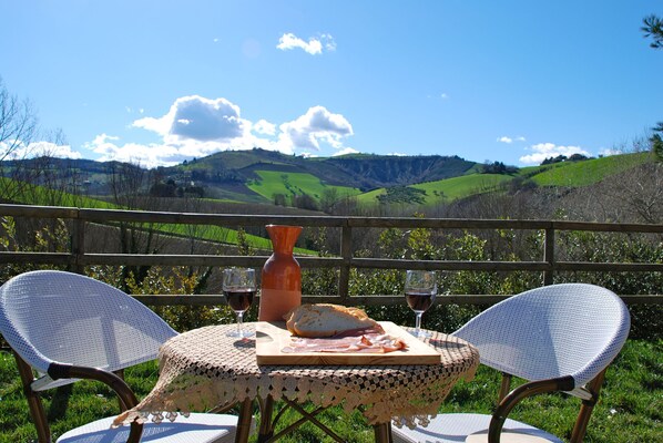 A table with a woven tablecloth, two chairs, a loaf of bread, a bottle, and two glasses of red wine, set against a scenic hillside view.