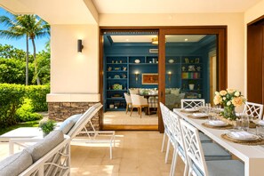 An outdoor dining area with a stone wall, white furniture, and a view into a room with a blue wall and bookshelves.