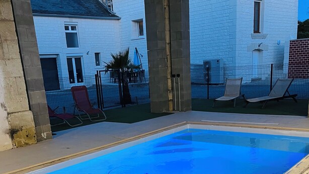 Une piscine avec vue sur un bâtiment en briques blanches et un patio clos, aménagé avec du mobilier d’extérieur.