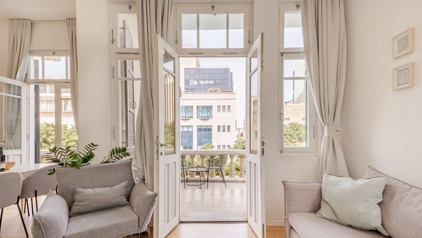 Ein modernes Wohnzimmer mit Sofa, Sessel und Esstisch. Ein Balkon bietet Blick auf Gebäude und Grünflächen.