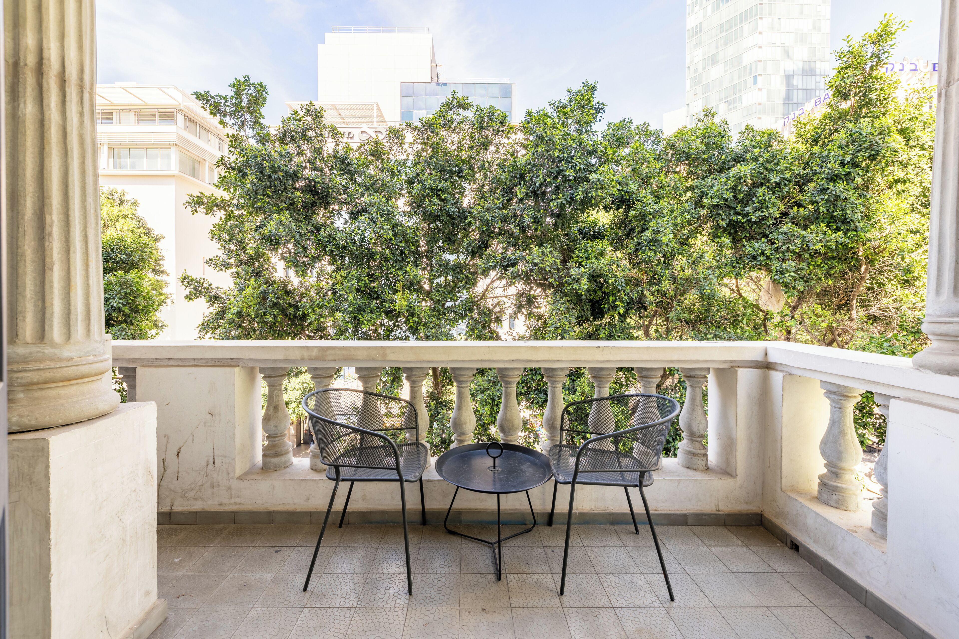 A balcony with two chairs and a table, overlooking a garden with trees and buildings in the background.