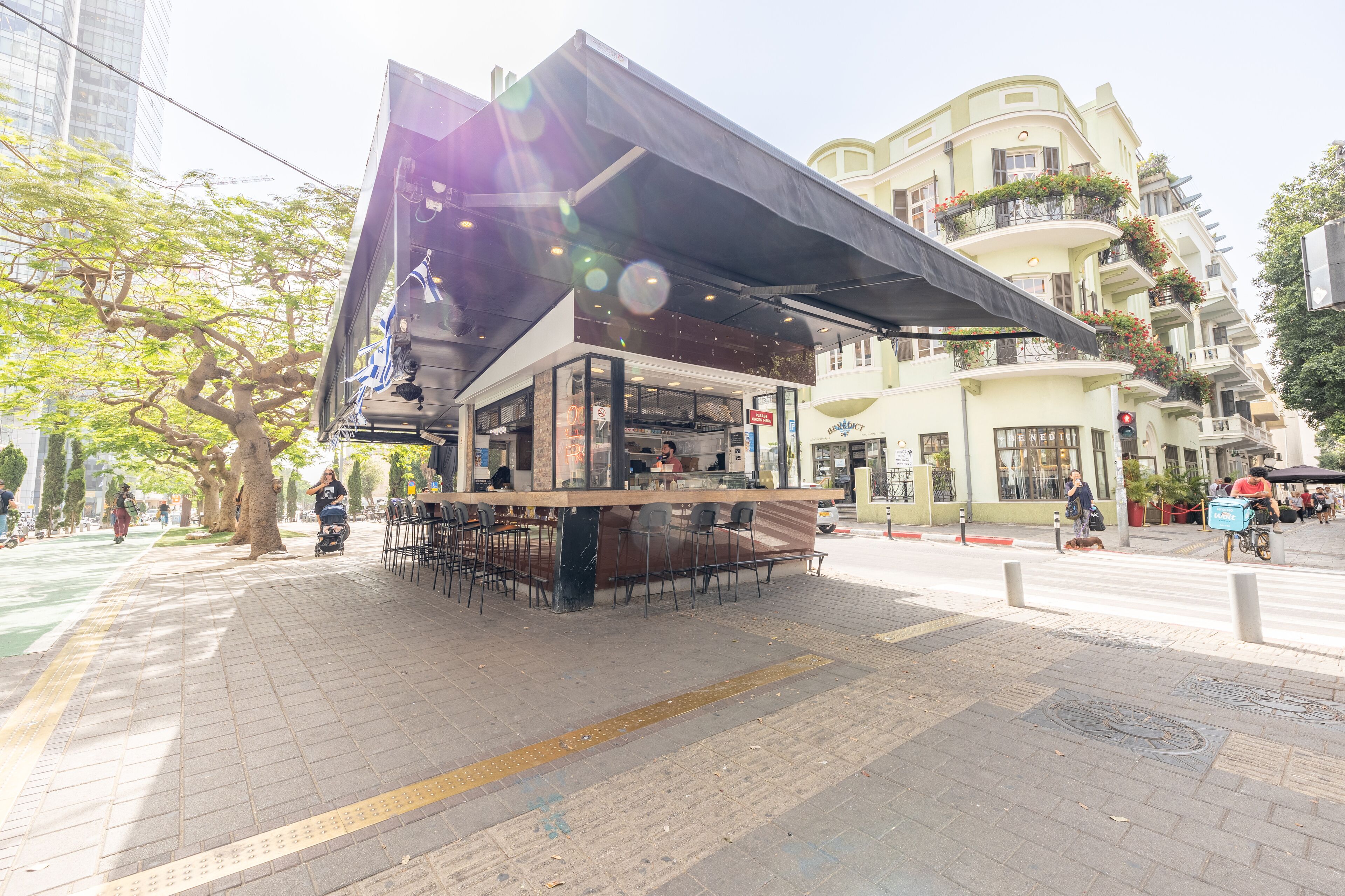 Un moderno café al aire libre con zona de estar cubierta, entrada visible y vista a la calle con peatones y vehículos.