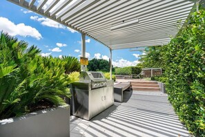 Enjoy al fresco dining under a shaded pergola, surrounded by lush greenery and a stainless steel barbecue grill.