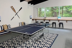 A ping pong table in a room with a patterned rug, a window with a view of trees, and a shelf with items.