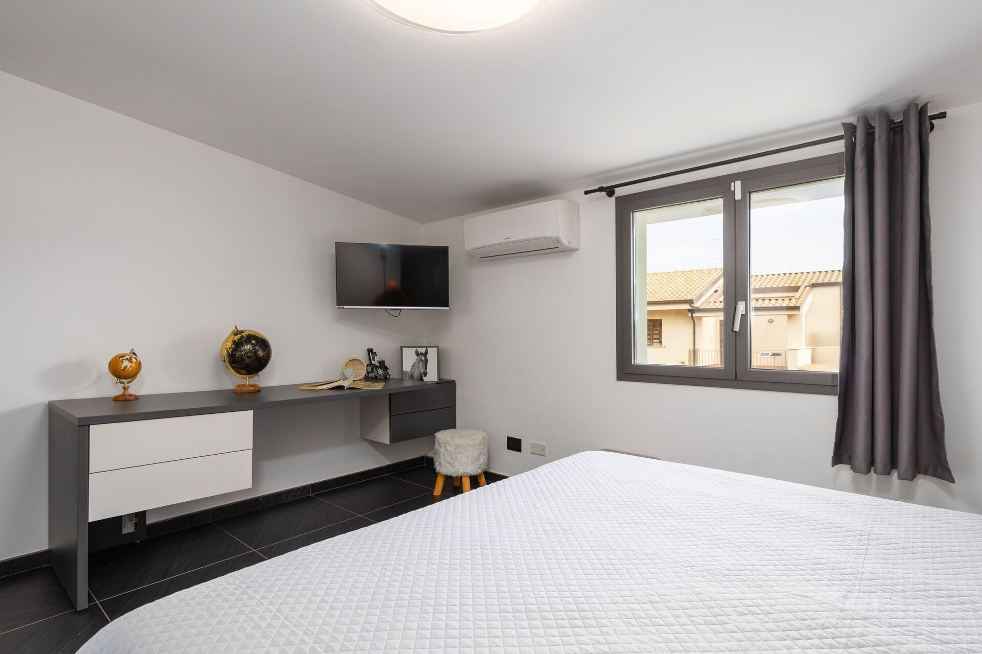 Une chambre à coucher moderne avec un lit, un bureau doté d’un téléviseur, un globe terrestre et une fenêtre avec des rideaux.