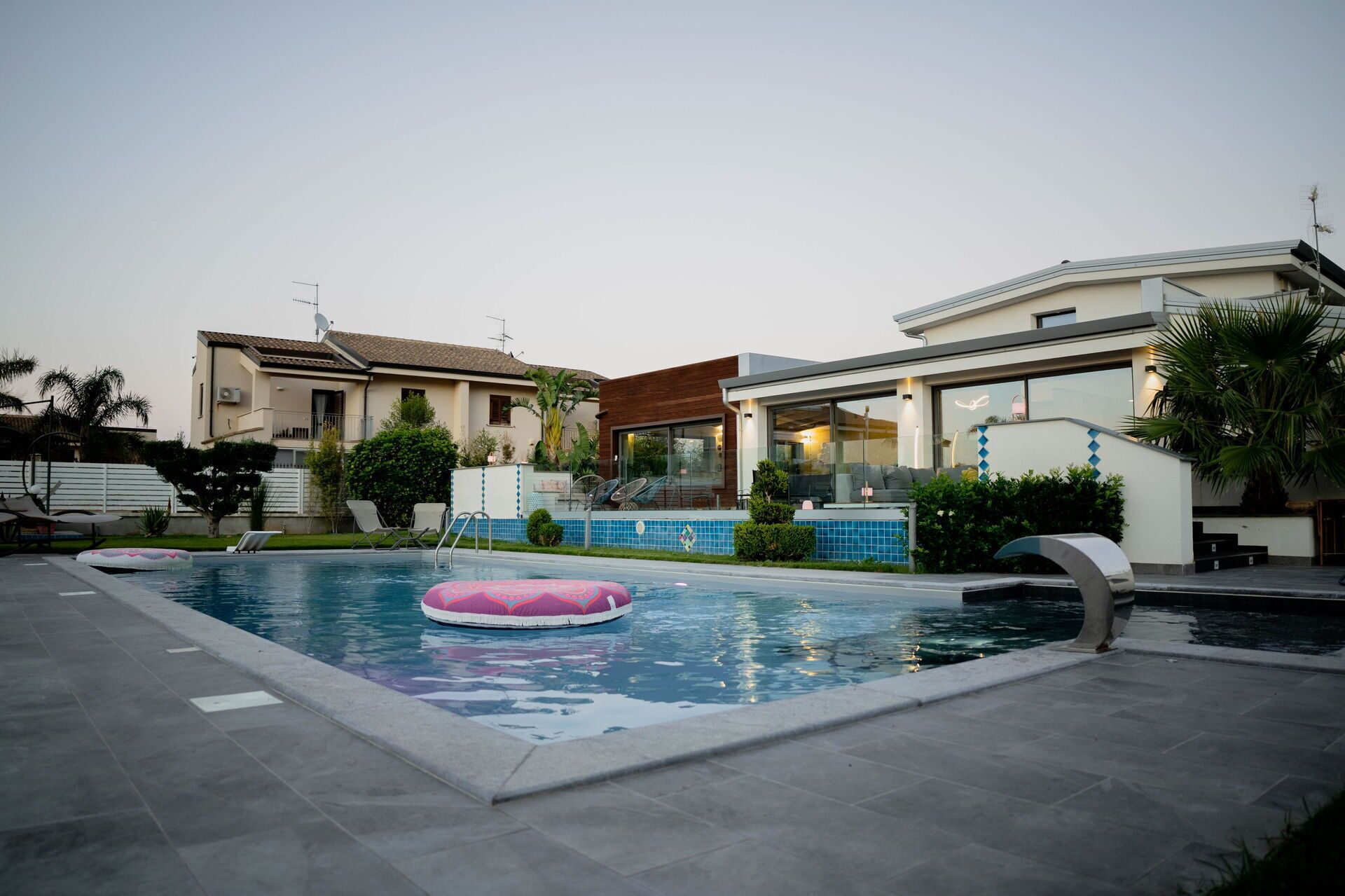 Une piscine avec un flotteur rose, entourée d’une terrasse aménagée avec des chaises et un petit jardin.