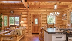 A log cabin interior with a wooden dining area, a sofa, a kitchen with a marble countertop, and a chandelier. - Bear Kub Kabin + Games + 1 Block to Snow Summit! (Big Bear Lake)