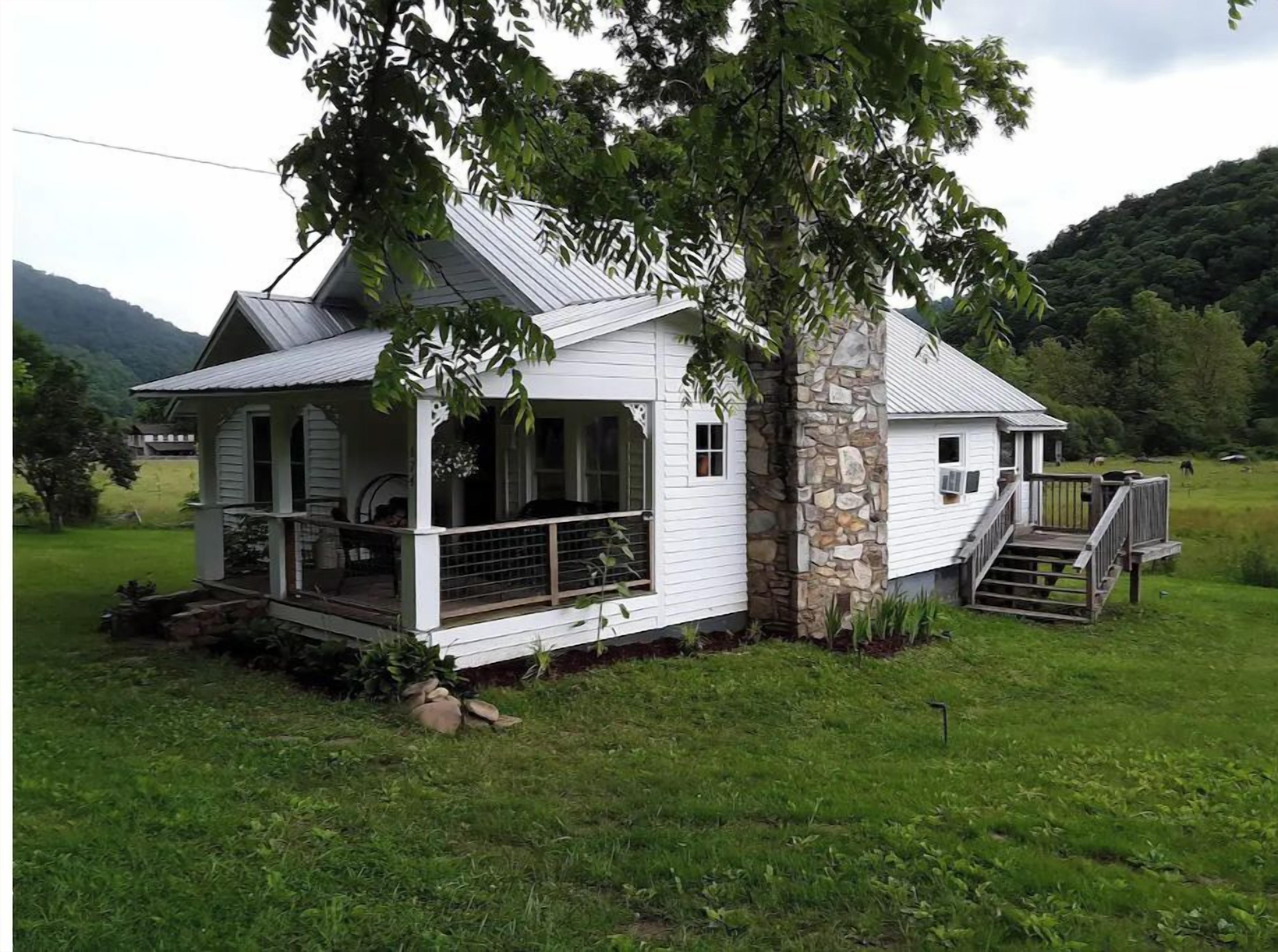 A white wooden house with a porch and stone foundation, surrounded by a grassy field and trees.