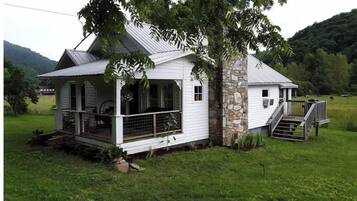 A white wooden house with a porch and stone foundation, surrounded by a grassy field and trees.
