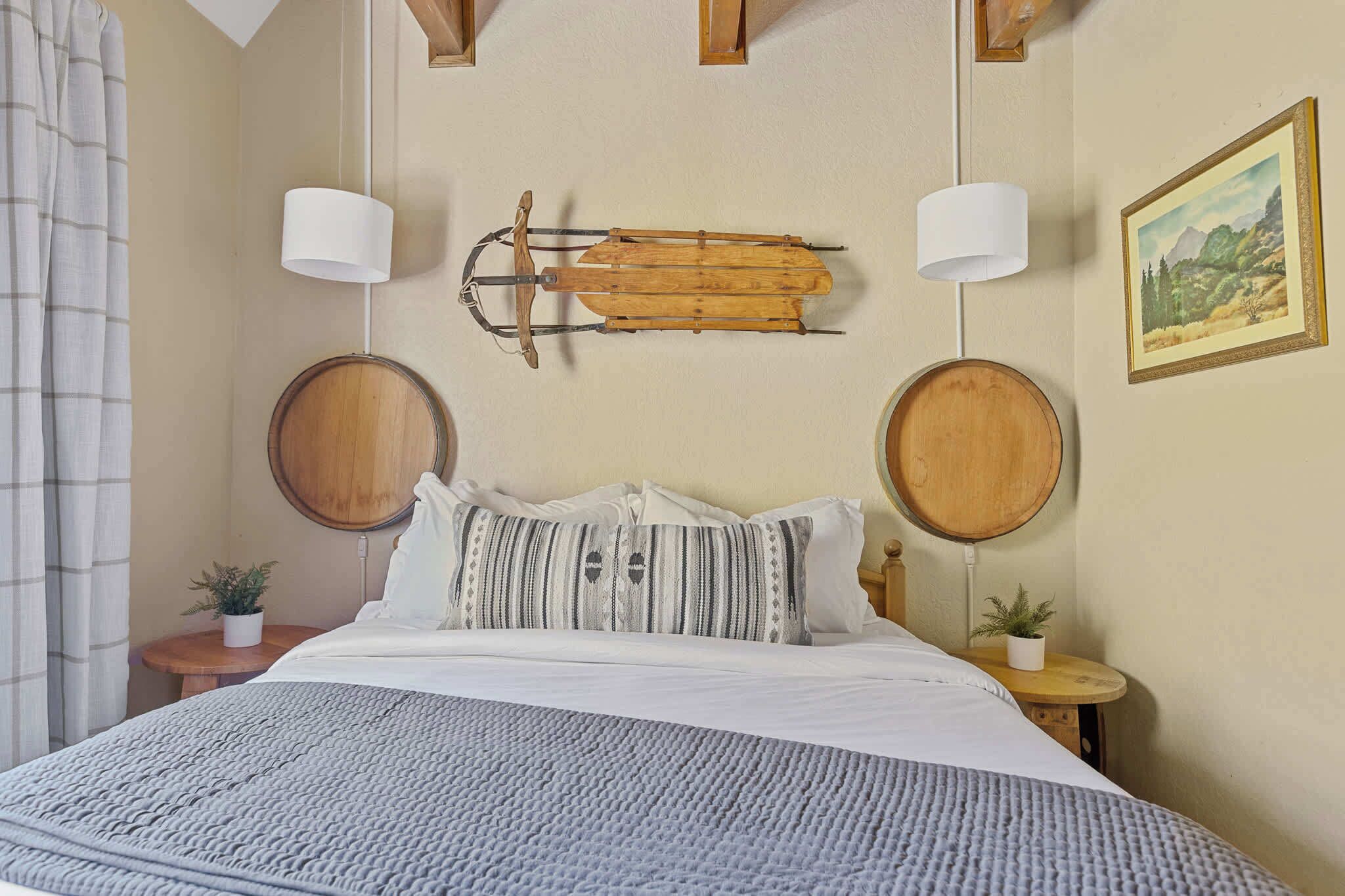 Un dormitorio con cama, dos mesitas de noche, un tronco de madera montado en la pared, dos lámparas de pared y un cuadro con paisaje enmarco.