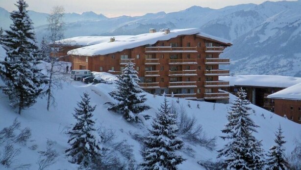 Un edificio de varios pisos con balcones, rodeado de árboles y montañas cubiertas de nieve.
