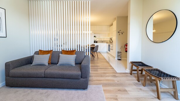 Un salón moderno con un sofá gris, taburetes de madera, un separador blanco y un espejo redondo en la pared.