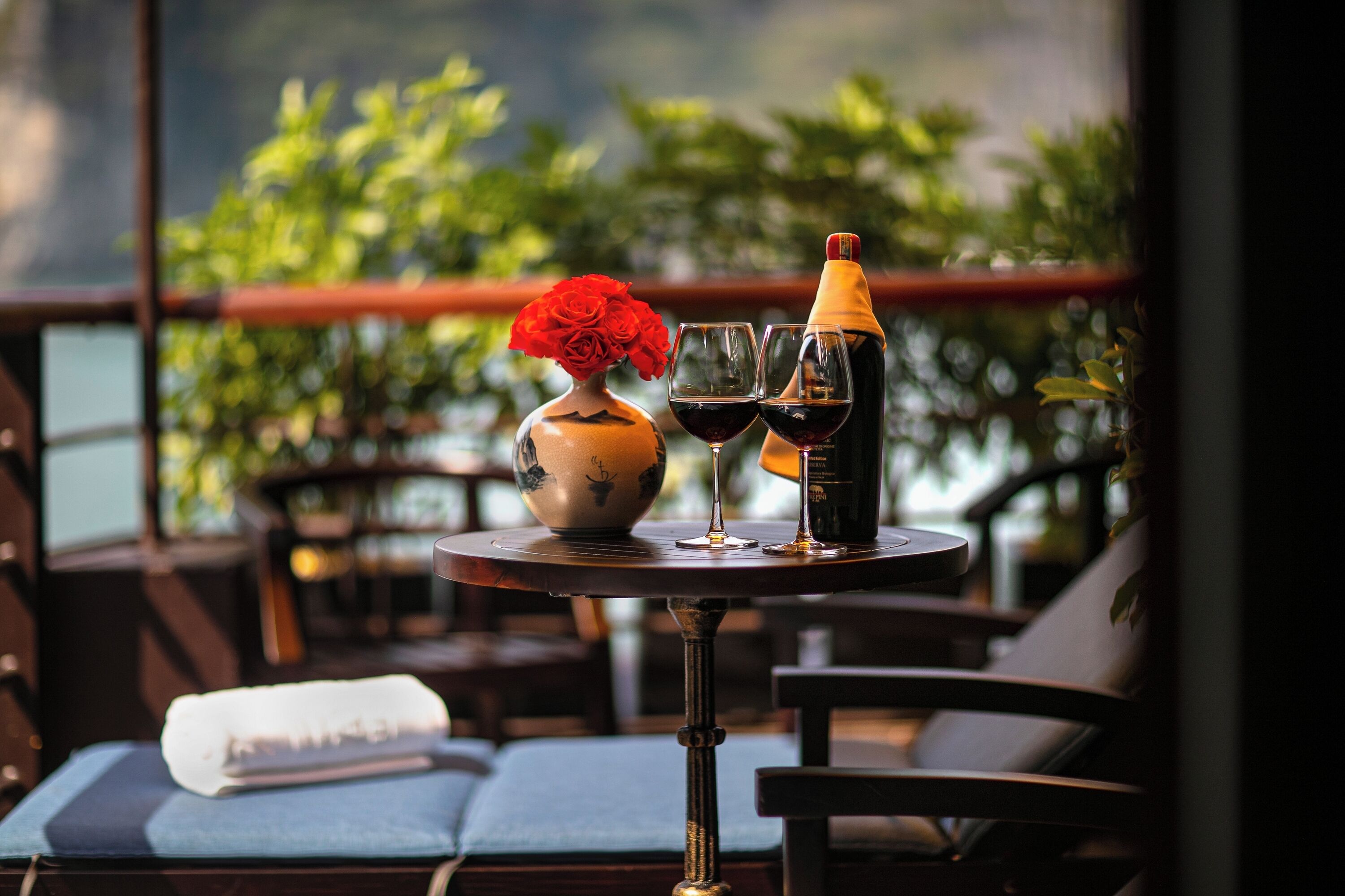 A table with a vase of red roses, two wine glasses, and a bottle of wine on a balcony with chairs and a view of greenery.
