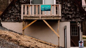 A wooden cabin with a balcony, a door, and a sign indicating a river. - Muskie-Lounge River Retreat! Secluded getaway.  (Buckhannon)