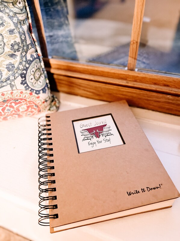 Un cuaderno de espiral con una etiqueta que dice 'Libro de visitas' y '¡Disfrute de su estancia!' colocado sobre una superficie junto a una ventana.