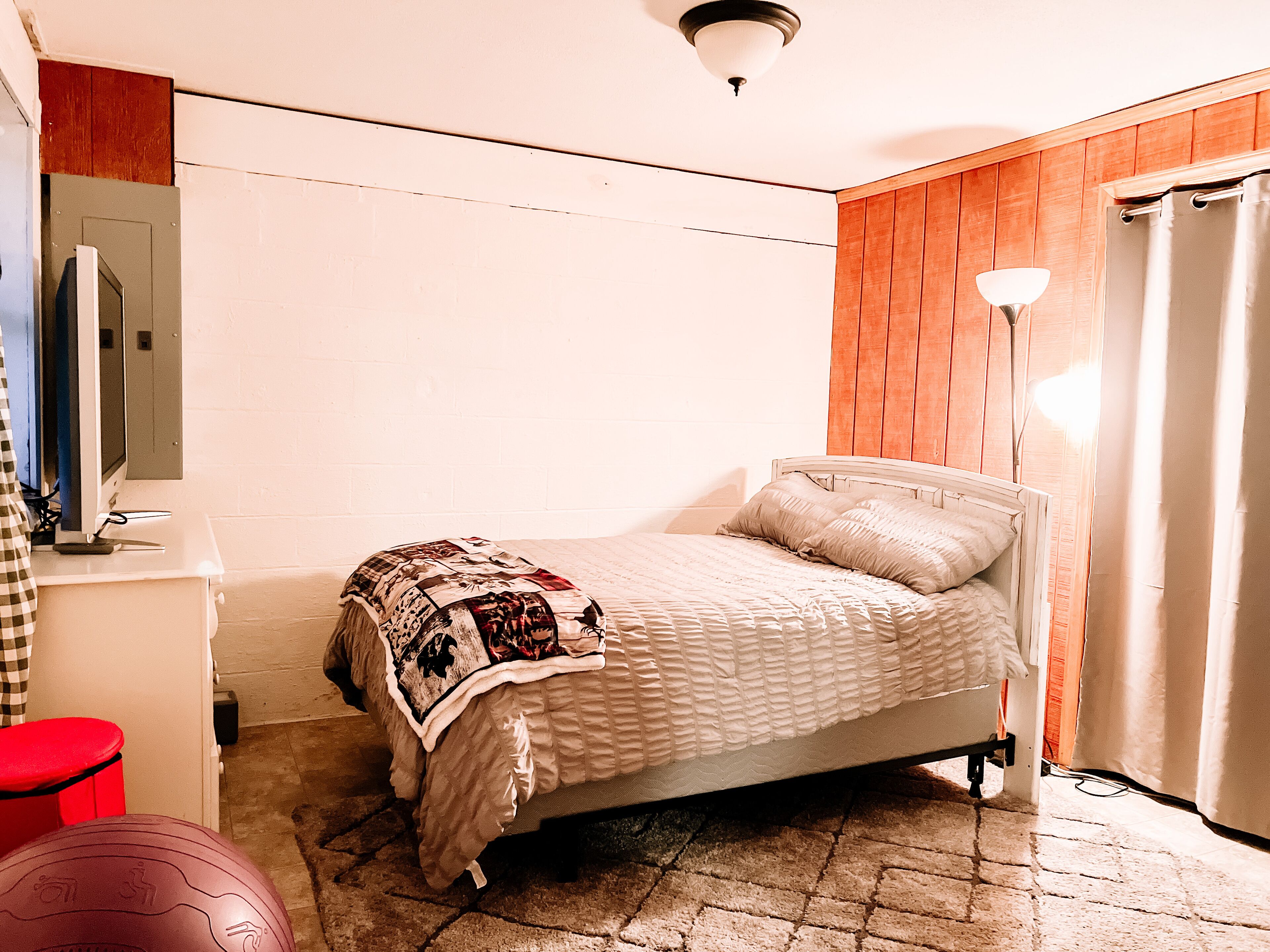 Un dormitorio con una cama, un escritorio, una silla, una lámpara y un taburete rojo.