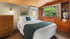 Une chambre avec un plafond en bois, un lit avec une literie blanche et un plaid vert, une commode et une fenêtre donnant sur un paysage verdoyant.