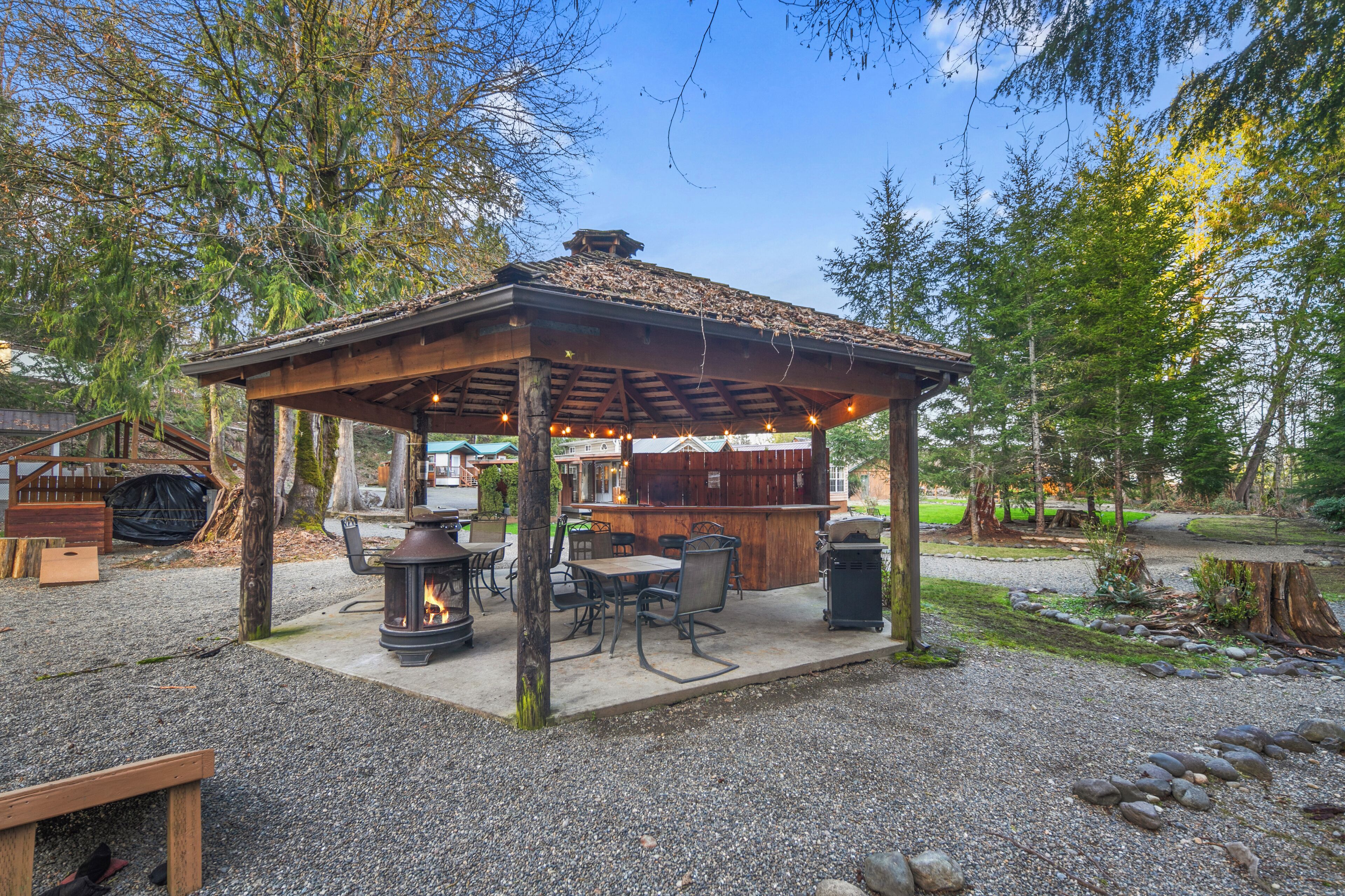 A gazebo with a fire pit and outdoor seating, surrounded by trees and a gravel path.