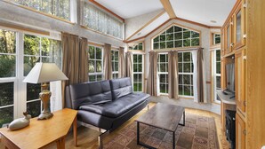 Un salón amplio con un sofá de cuero, una mesa de centro de madera y un tapete estampado. Grandes ventanas con cortinas ofrecen vistas a la vegetación exterior.