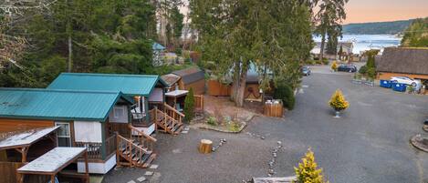 A rustic cabin area with multiple wooden structures, a gravel driveway, and a view of the water in the background.