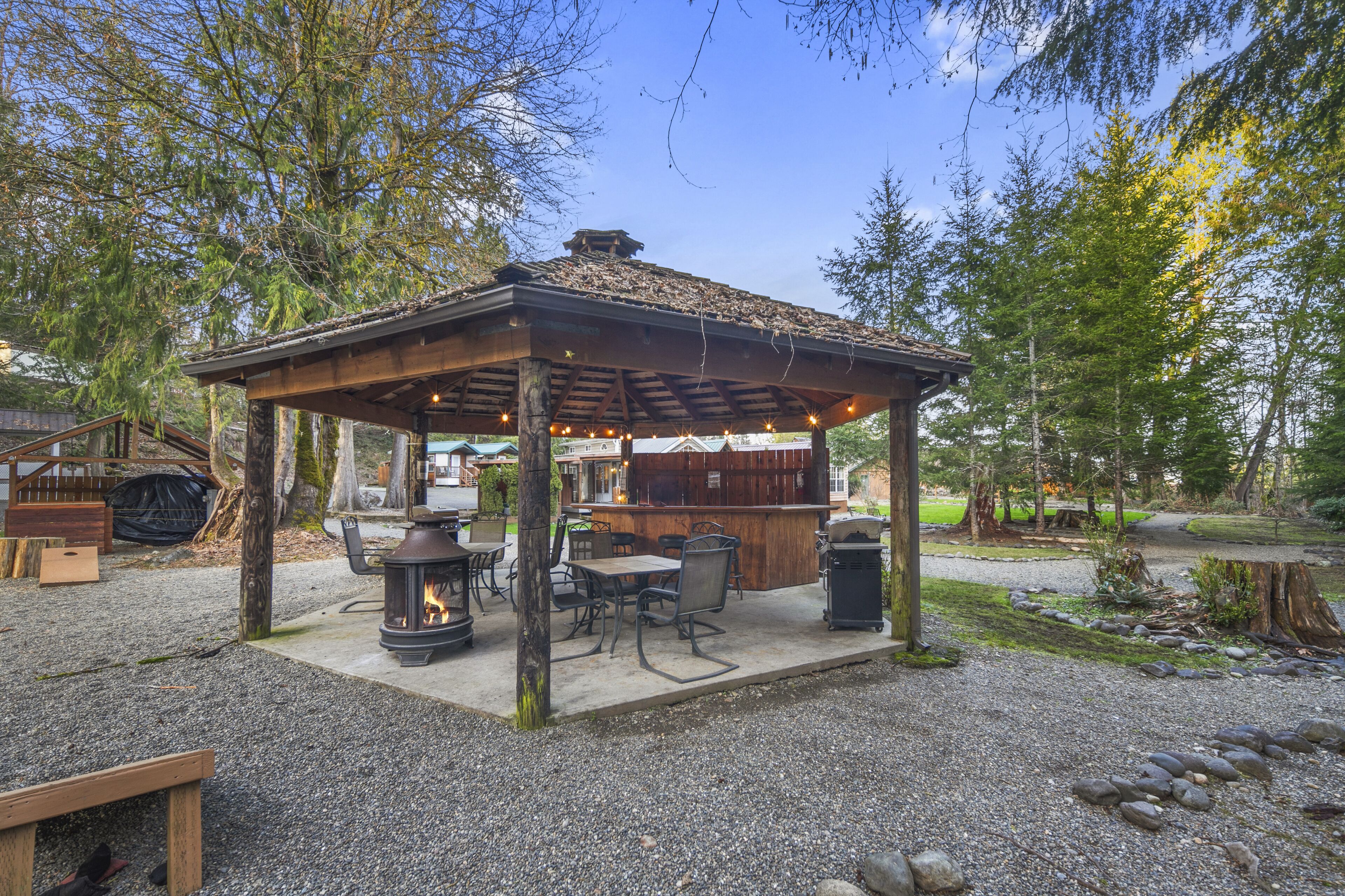 A gazebo with a fire pit and outdoor seating, surrounded by trees and a gravel path.