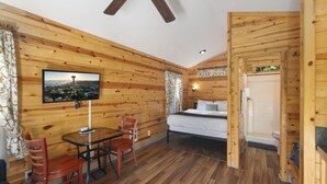 Eine Holzhütte mit einem Bett, einem Esstisch mit Stühlen, einem an der Wand befestigten Fernseher und einem durch eine offene Tür einsehbaren Badezimmer.