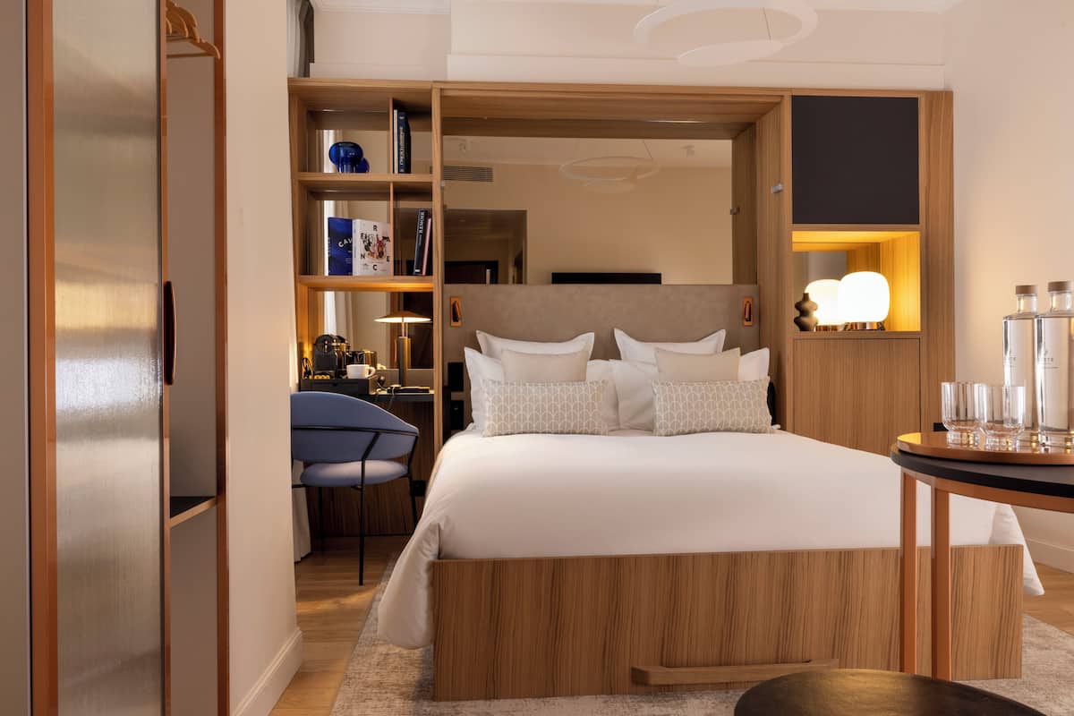 Una habitación de hotel moderna con una cama grande, cabecero de madera, un escritorio con silla, una estantería y una mesita de noche con vasos.