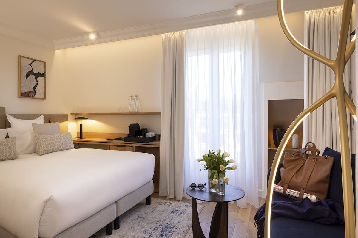 Habitación de hotel con una cama grande, una mesita de noche con una lámpara, una pequeña mesa con un jarrón de flores y una silla con un bolso de mano.