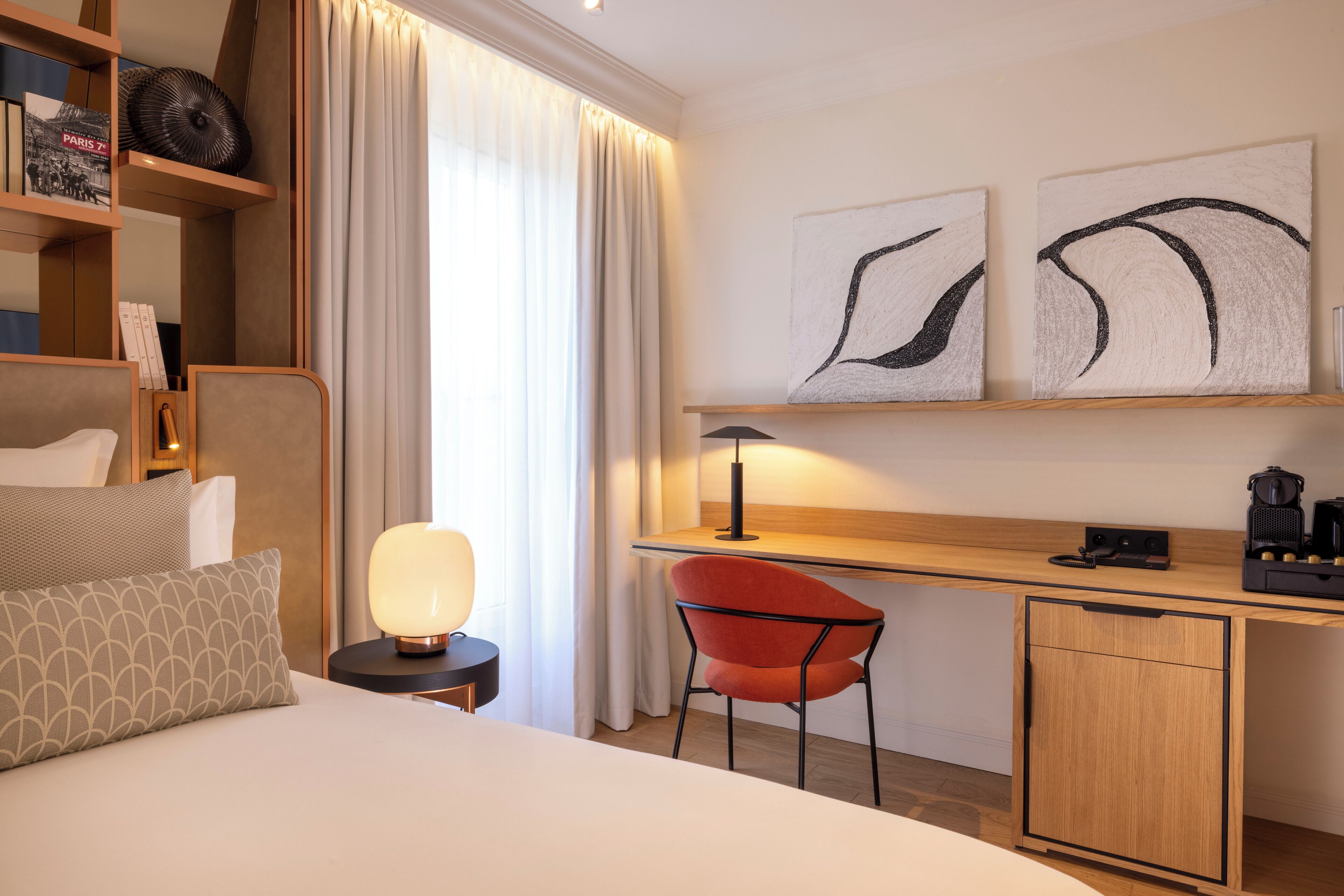 Une chambre d’hôtel comprenant un lit, un bureau avec une chaise, une lampe, une machine à café et des œuvres d’art accrochées au mur.