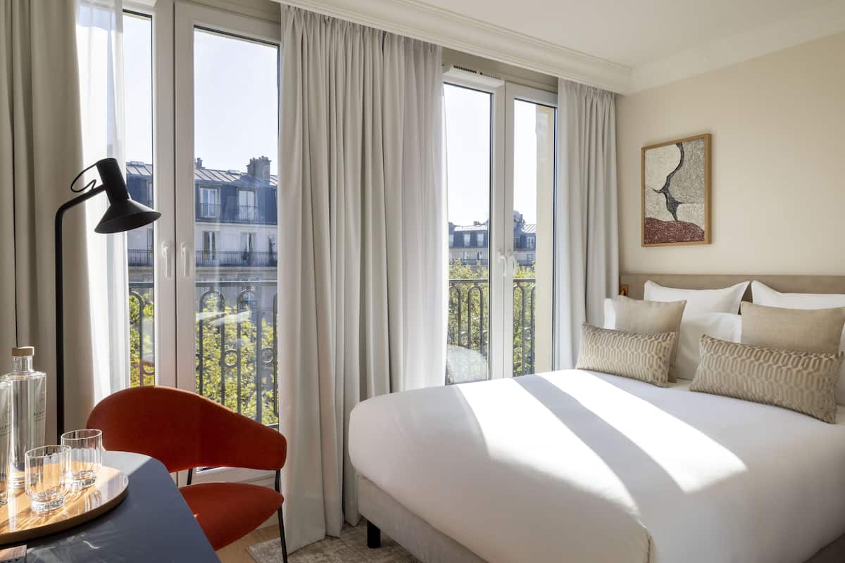 Una habitación de hotel con una cama grande, una silla, una mesa con vasos y una botella, y vistas a edificios a través de amplios ventanales.