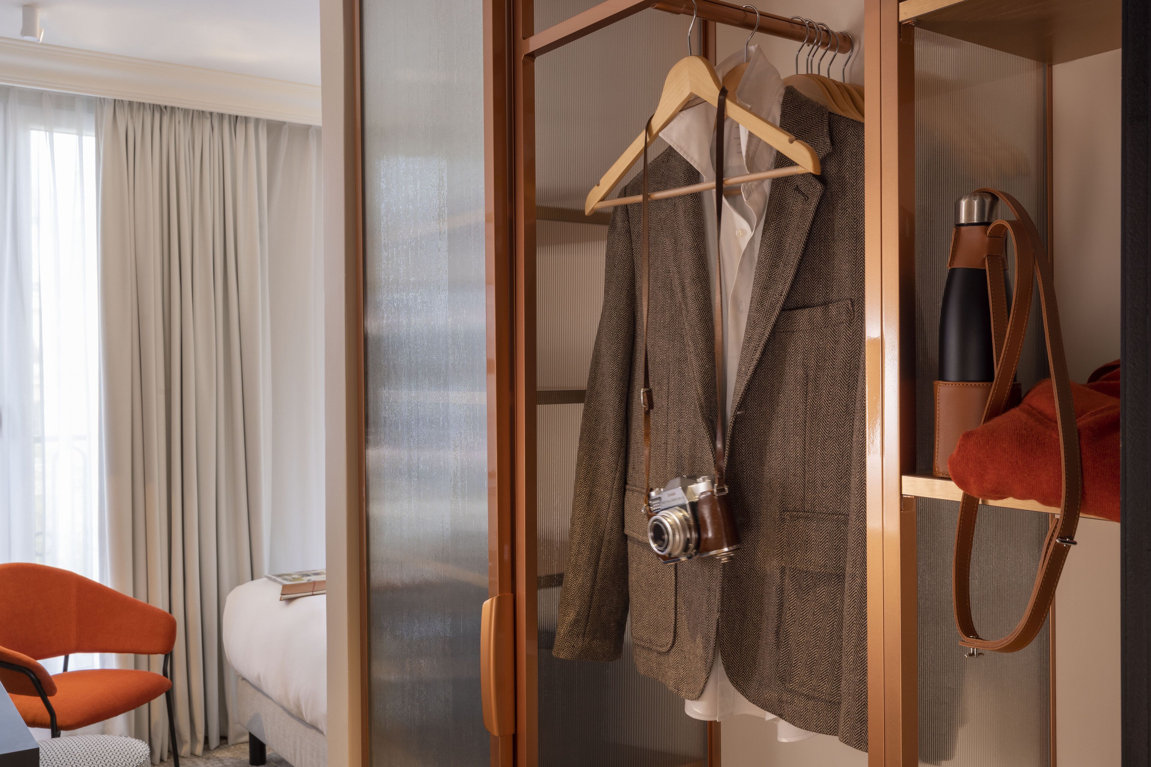 A hotel room with a bed, a chair, a wardrobe with hangers, and a camera.