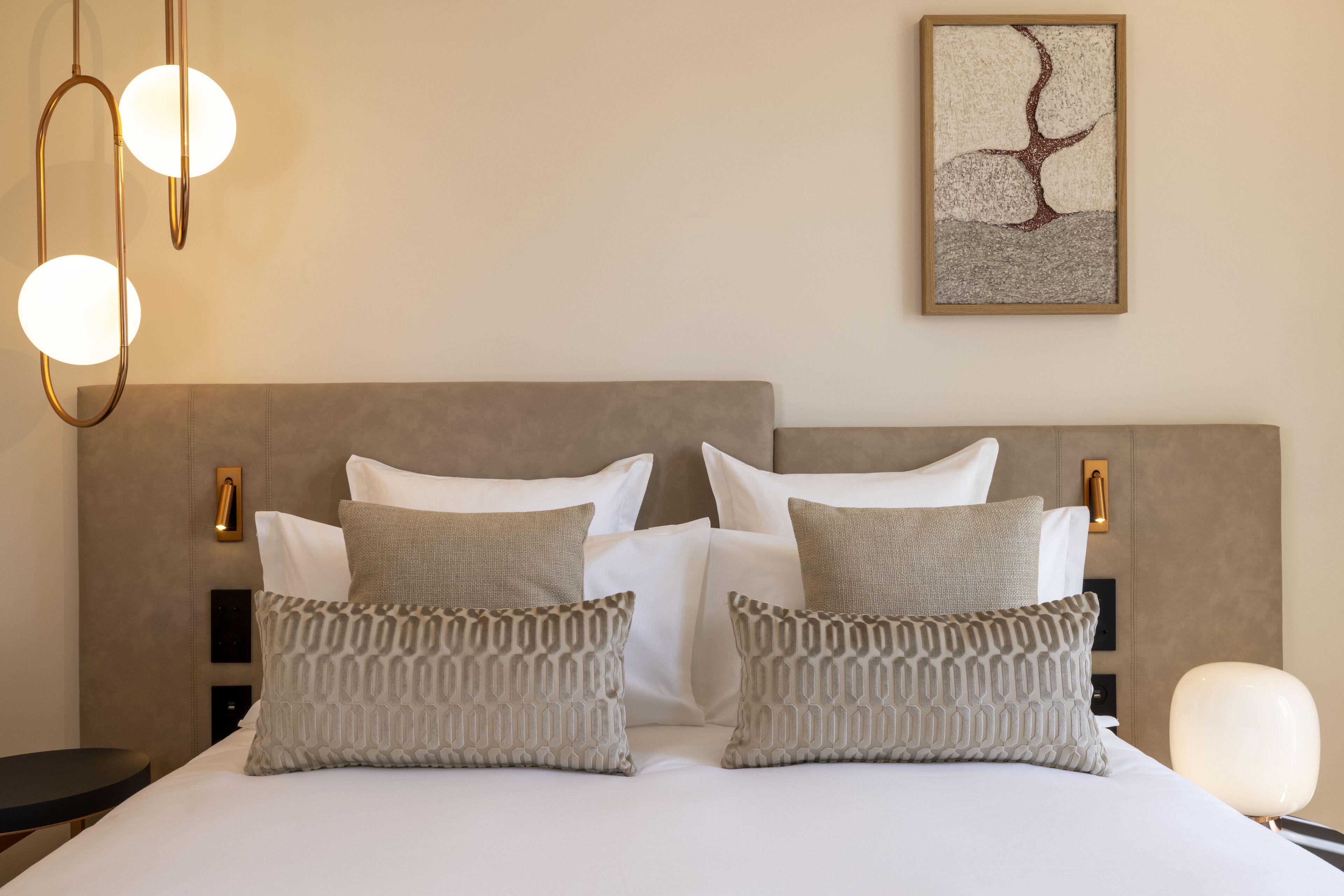 Una cama bien tendida con cabecera gris, dos almohadas estampadas y un cuadro abstracto colgado en la pared.