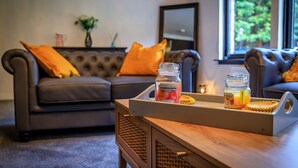 A living room with a dark brown sofa, two orange cushions, a wooden coffee table with a tray holding a candle and a jar of orange juice. - Opulent, relaxing home with hot tub (Fochabers)
