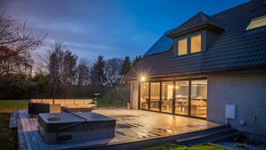 A modern house with a deck, hot tub, and glass doors leading to a living area. - Opulent, relaxing home with hot tub (Fochabers)