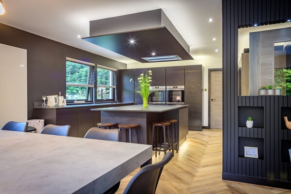 A modern kitchen with a central island, bar stools, and a dining area with a large table. - Opulent, relaxing home with hot tub (Fochabers)