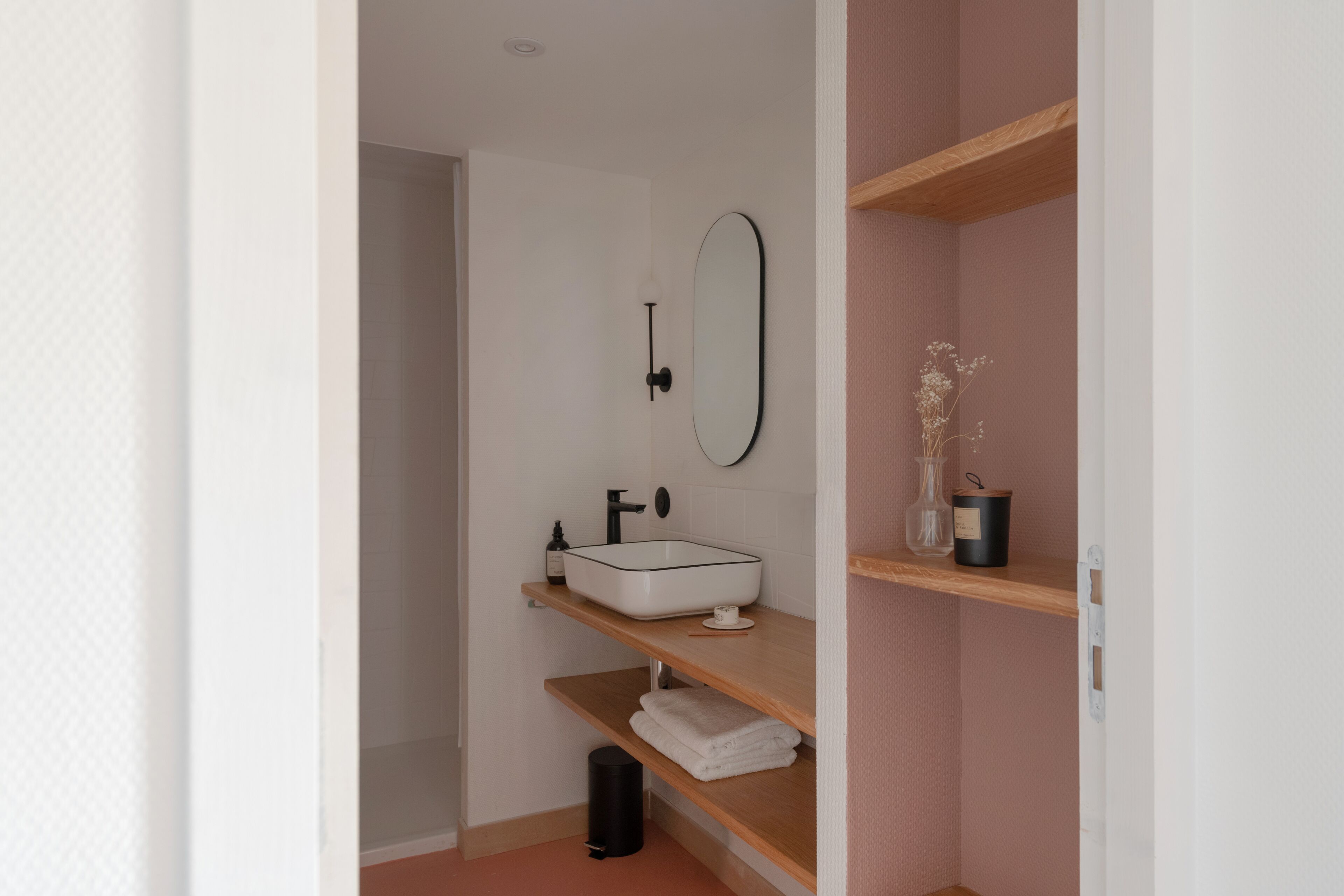 Un bagno moderno con un lavandino bianco, un rubinetto nero e uno specchio. Si vedono scaffali in legno con un vaso e una candela.