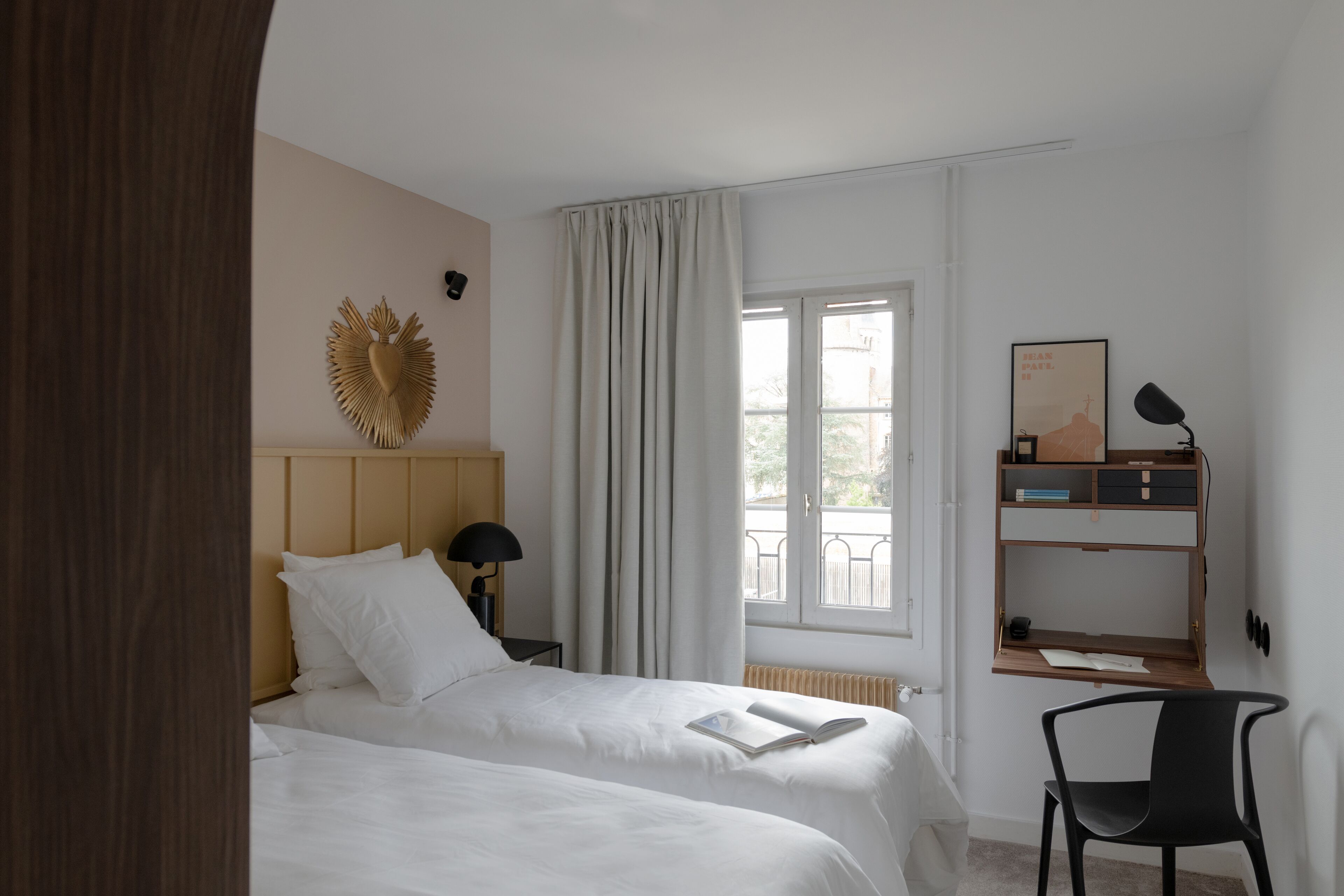 A hotel room with two beds, a desk with a chair, a window with curtains, a wall-mounted shelf, and a decorative wall piece.