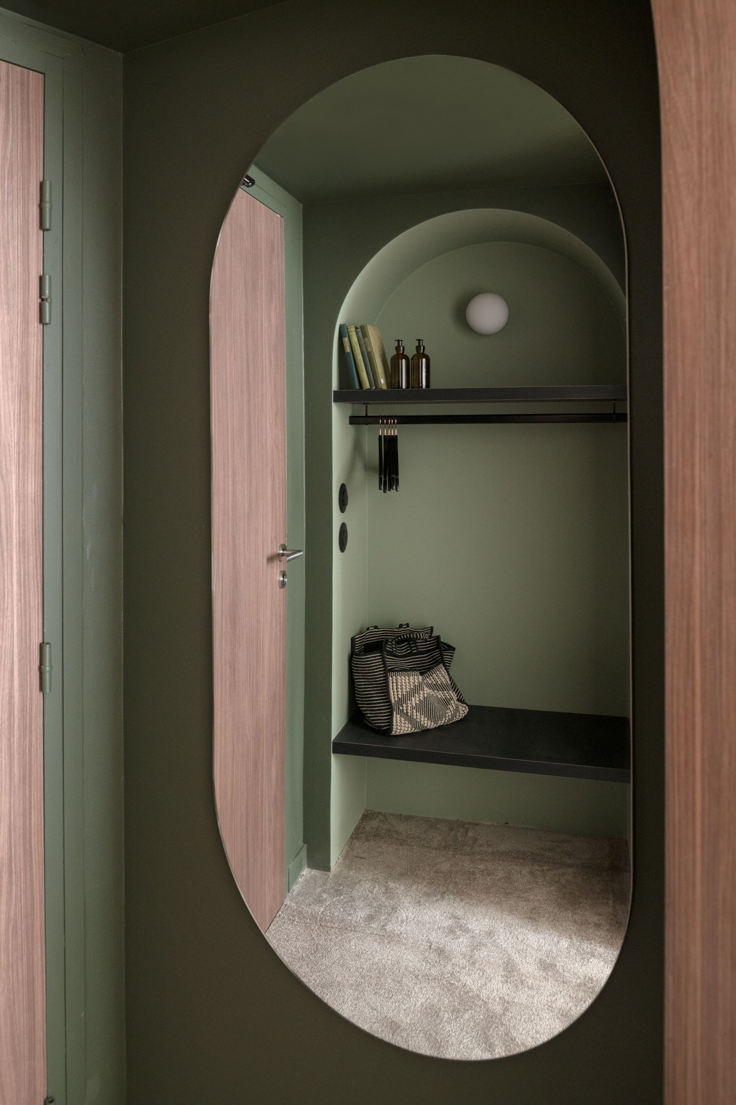 A room with a circular mirror, a green door, and a shelf with books and bottles.