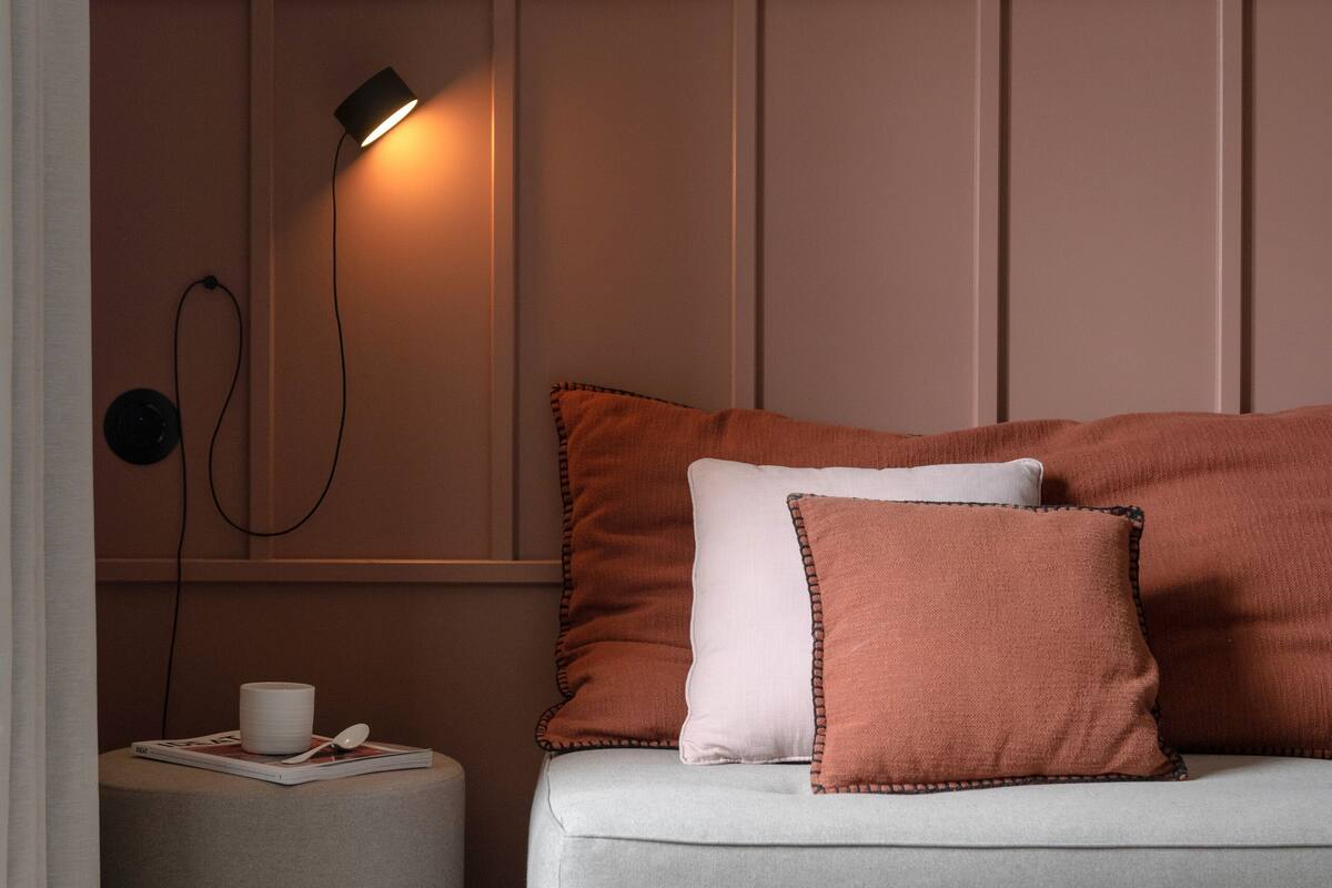 A cozy room with a sofa, pillows, a side table, a lamp, and a shelf with decorative items.