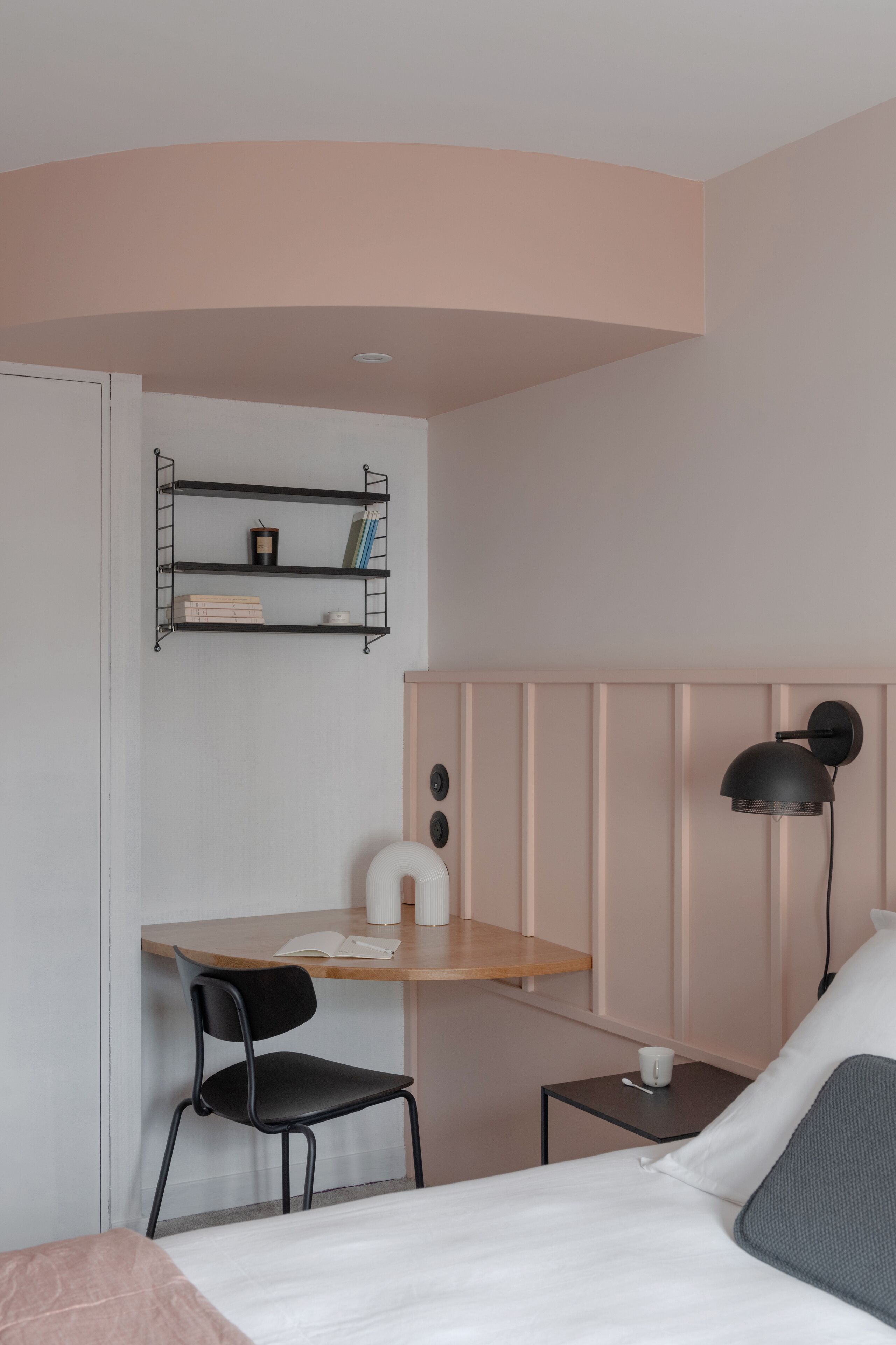 A bedroom with a bed, a desk with a chair, a shelf with books and a cup, and a lamp.