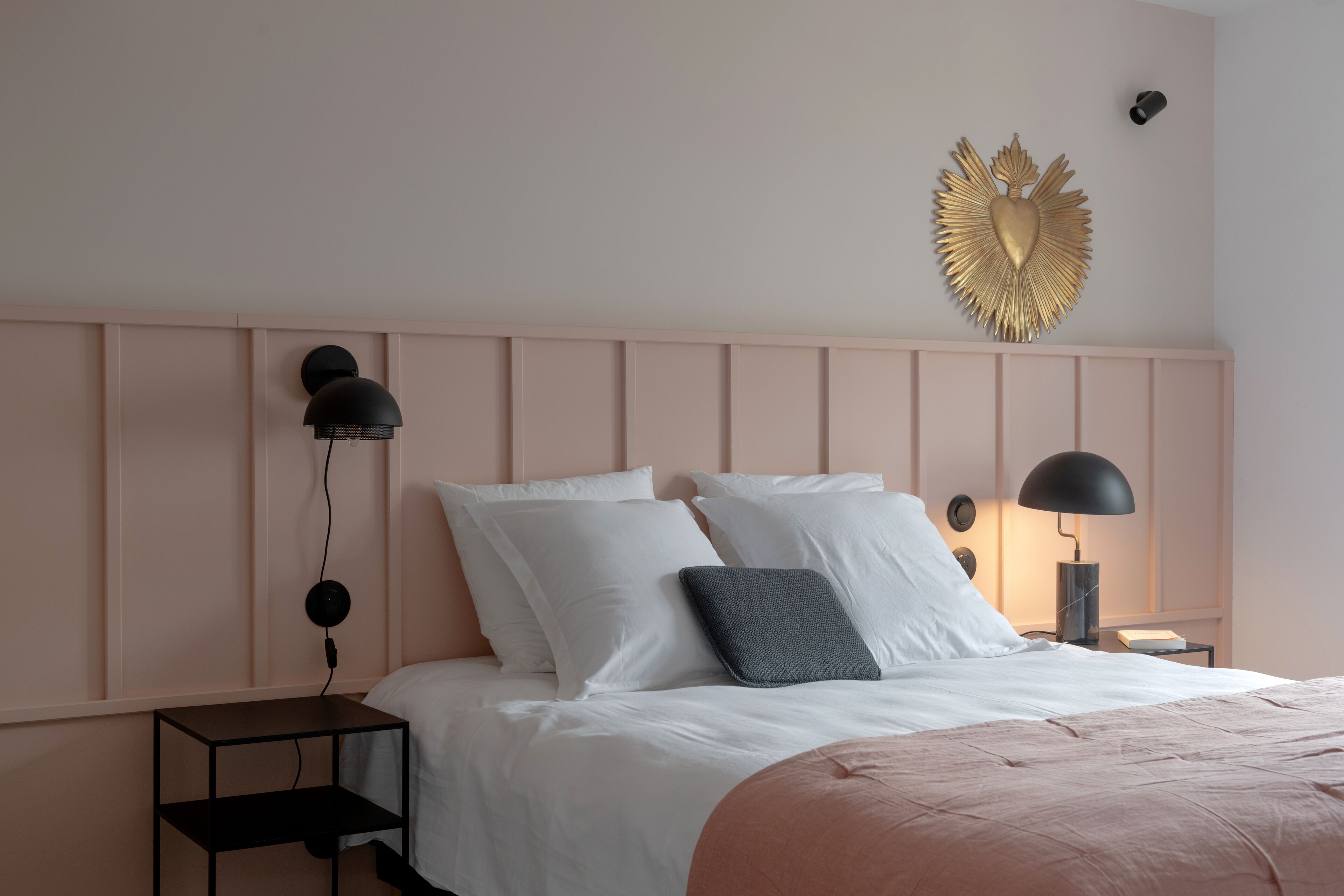 A neatly made bed with white bedding, a pink throw, and a gold decorative piece above it. There are two bedside tables, each with a lamp and a book.