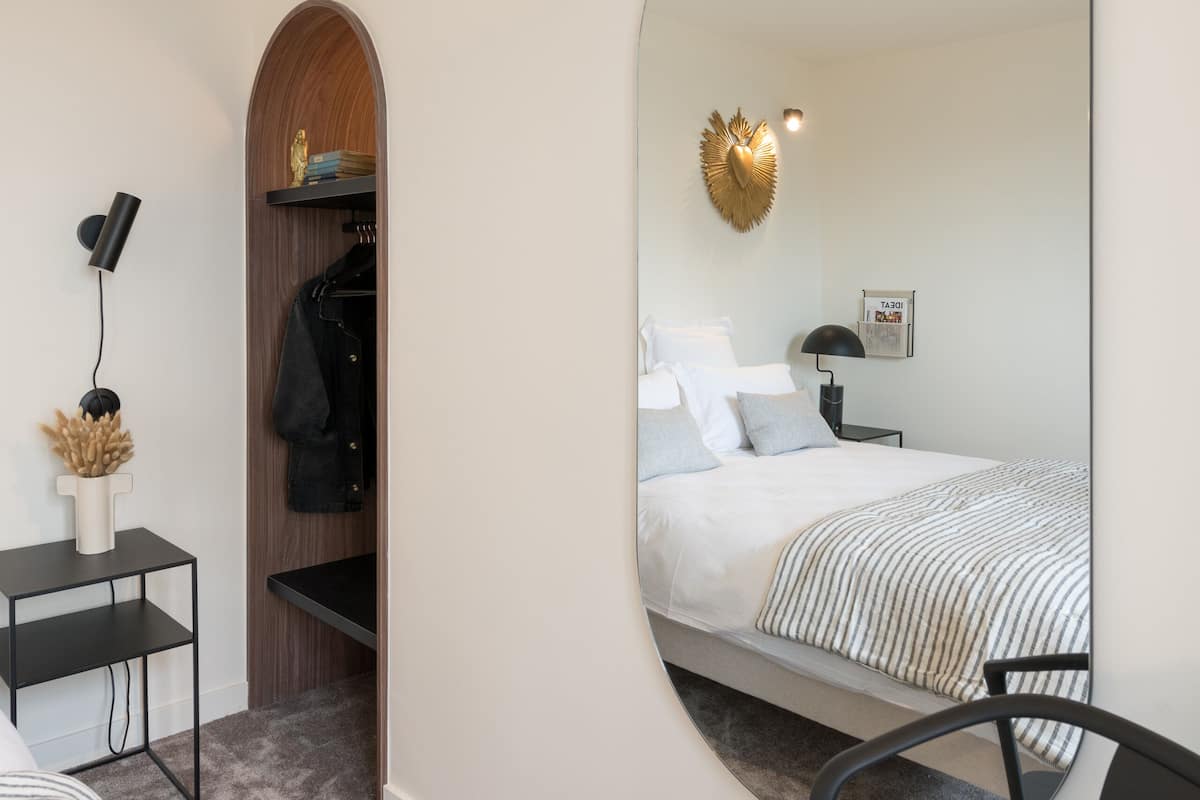 A modern bedroom with a large mirror, a bed with striped bedding, a desk with a lamp, and a coat rack.
