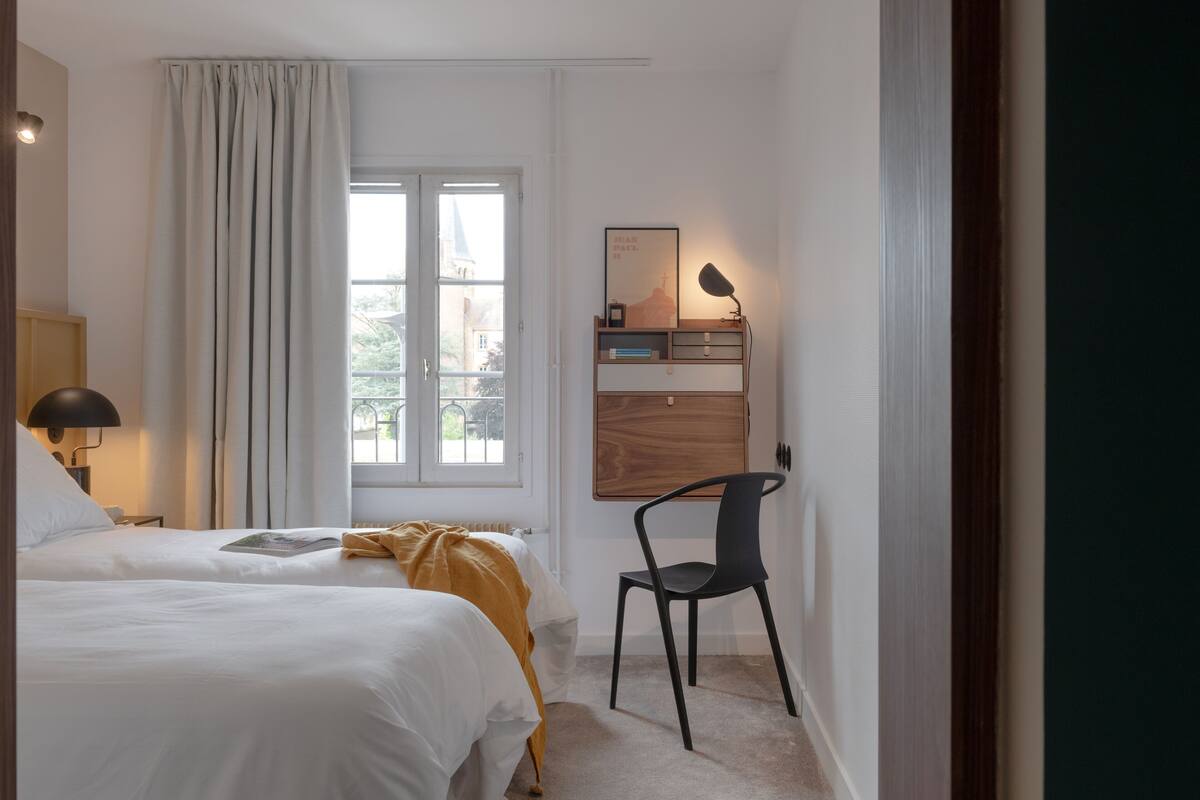 A hotel room with two beds, a desk with a lamp, a chair, and a window with curtains.