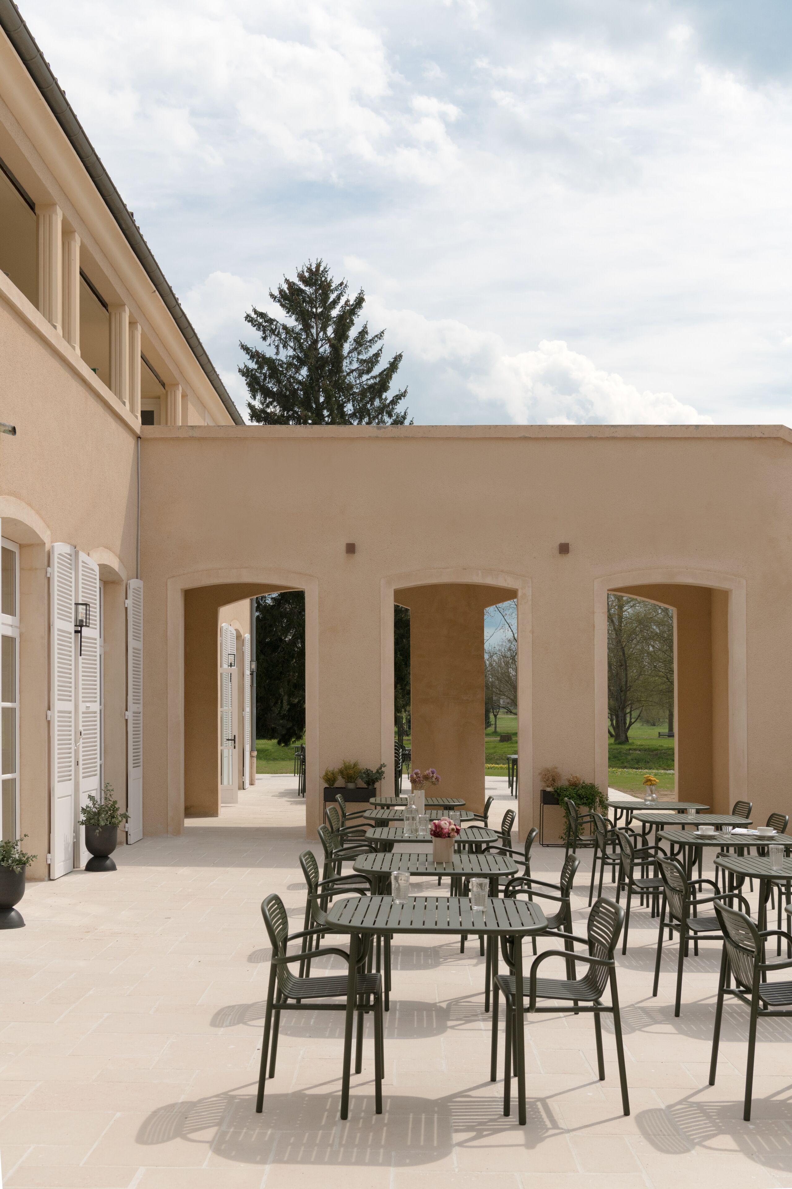 An outdoor patio with multiple tables and chairs, surrounded by arches and potted plants.