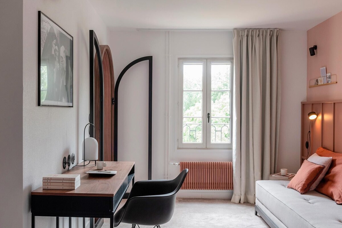 A room with a desk, chair, books, a radiator, a window with curtains, and a sofa with cushions.